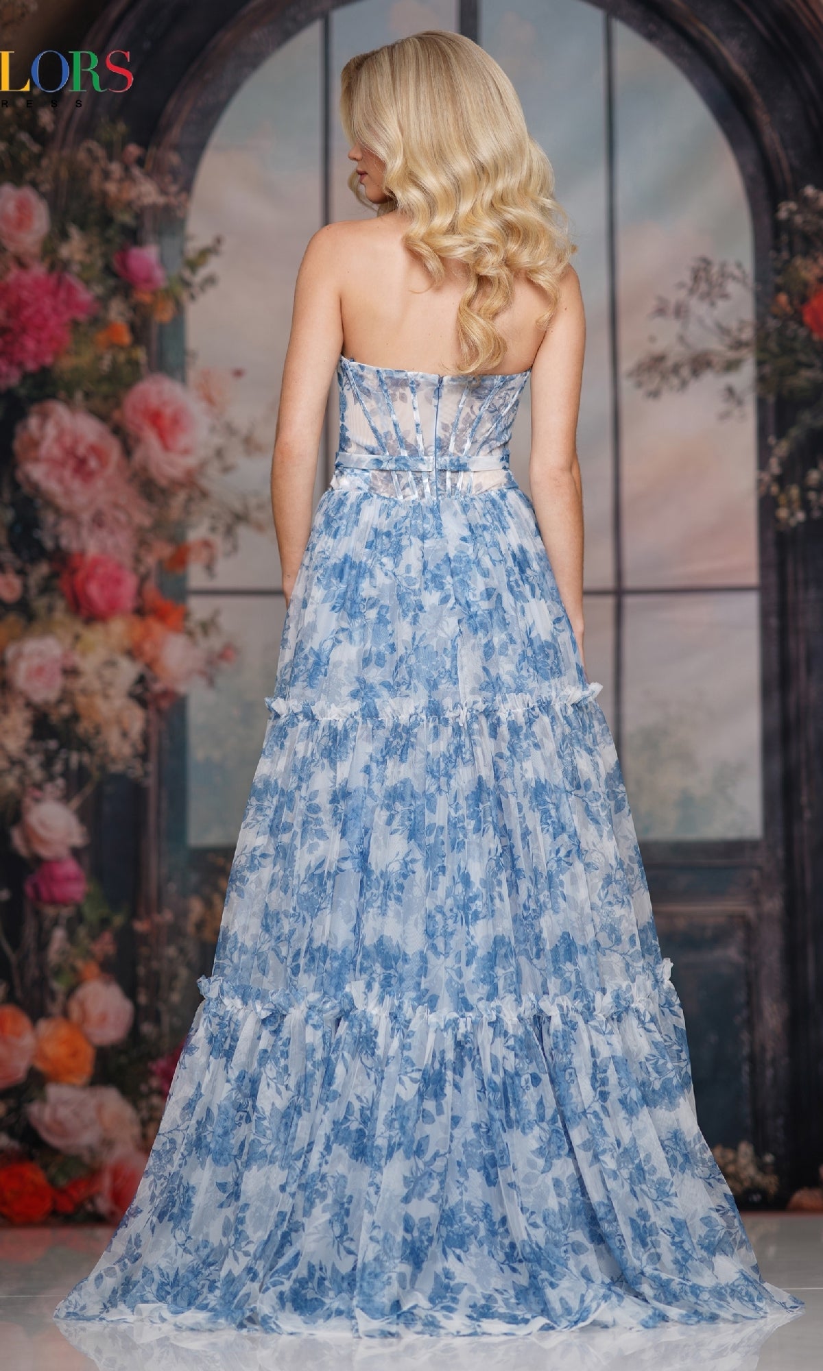 A woman with long blonde hair models the Strapless Long Print Prom Dress: Colors Dress 3810, standing with her back to the camera before an arched, flower-adorned window.